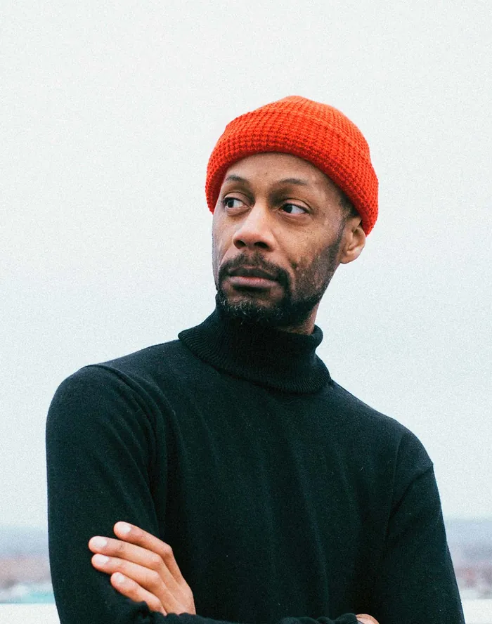 Portrait of a man in an orange beanie and dark turtleneck with his arms crossed.